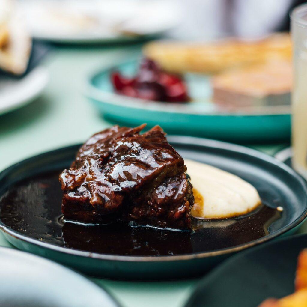 A close-up of a dark, glossy beef cheeks with polenta on a dark plate, surrounded by other blurred dishes in the background.