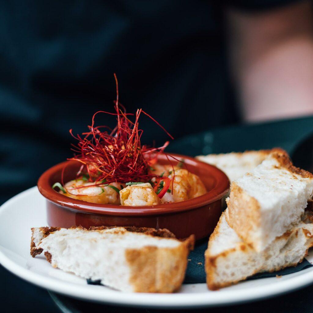 A small dish of cooked shrimp garnished with thin red chili threads is served on a white plate with several pieces of rustic bread.