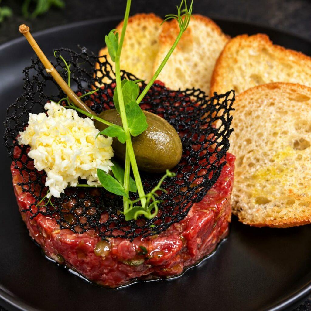 A serving of beef tartare topped with a black lace tuile, a green olive, minced egg, and pea shoots, served with three slices of toasted bread on a black plate.