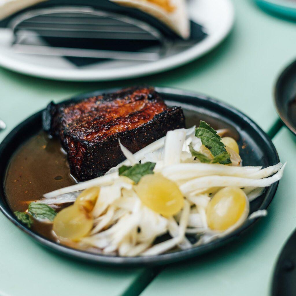 A plate with a piece of pork belly in sauce, served alongside a salad of shaved fennel, green grapes, and fresh mint leaves.