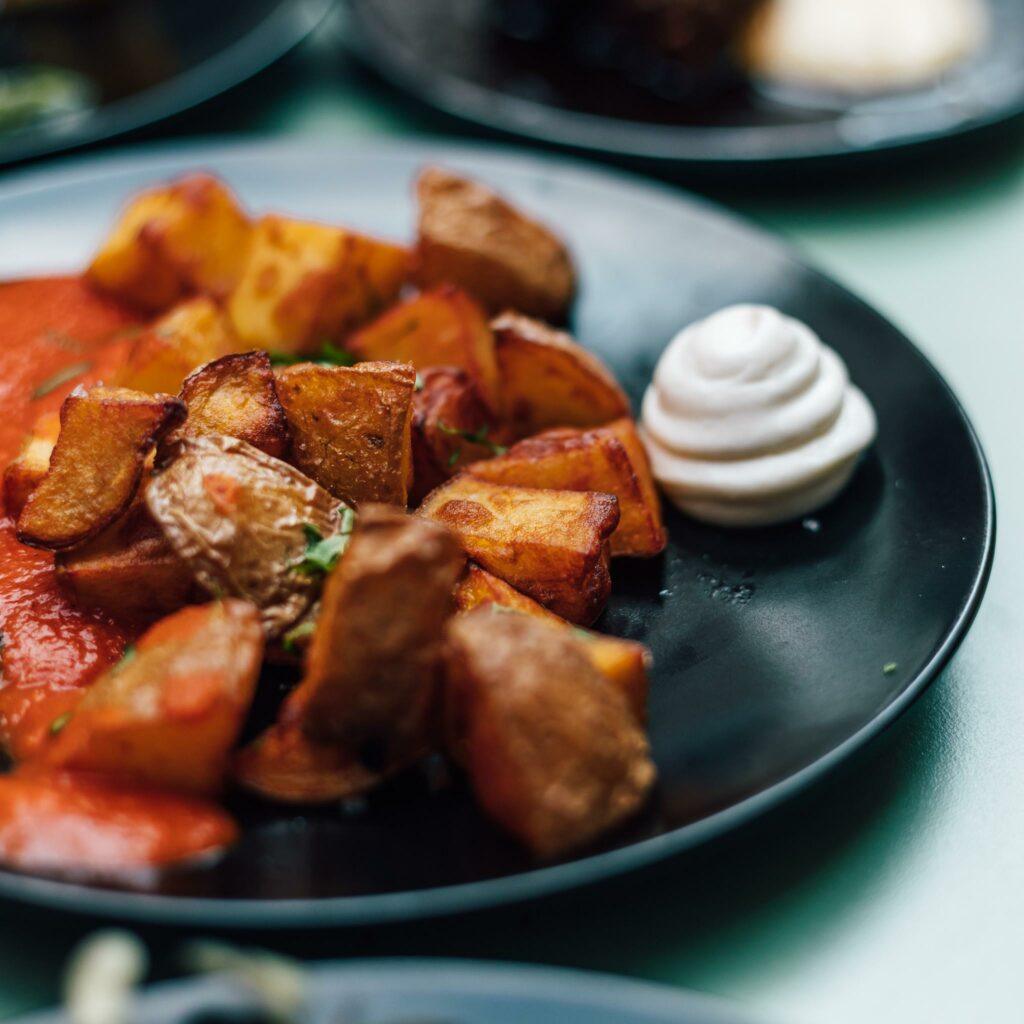 A close-up of a plate with crispy roasted potato cubes, a swirl of creamy white sauce, and a red sauce, served on a black plate. Patatas Bravas